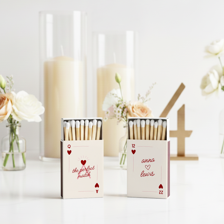 Two personalized wedding matchbox favors styled as playing cards, displayed on a white table with roses and tall cream candles in the background. One box reads “the perfect match” with heart symbols, and the other features custom names “anna ♥ lewis.”