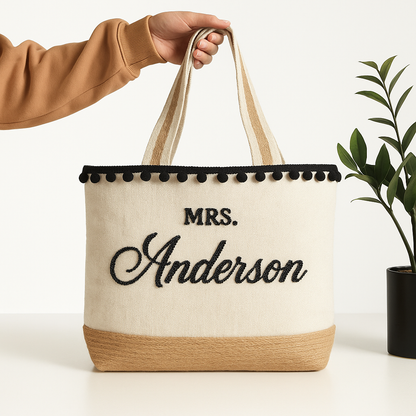 A hand holding a beige tote bag with the name "Mrs. Anderson" embroidered on it.