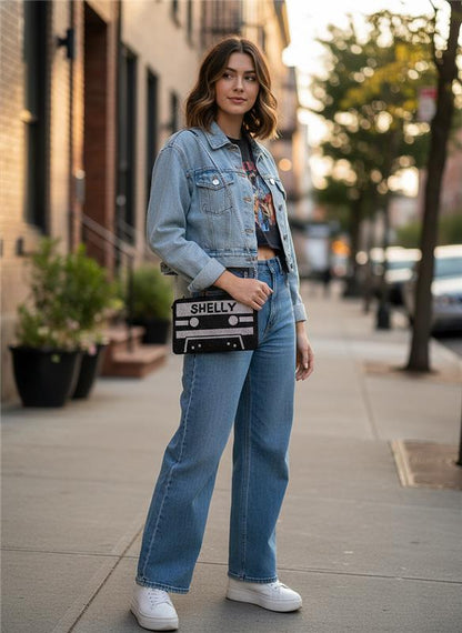 A young woman wearing a denim jacket, jeans, and sneakers is standing on a city sidewalk, holding a black and white bag with the word "SHELLY" printed on it.