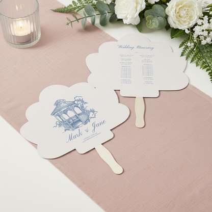 two fan-shaped wedding programs on a pink tablecloth, with a candle and greenery in the background.