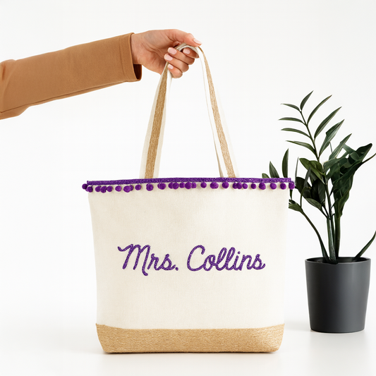 A hand holding a white tote bag with purple trim and the text "Mrs. Collins" embroidered on it, next to a potted plant.