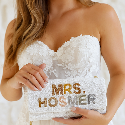 A woman wearing a white lace dress holds a white clutch purse with the text "Mrs. Hosmer" printed on it.