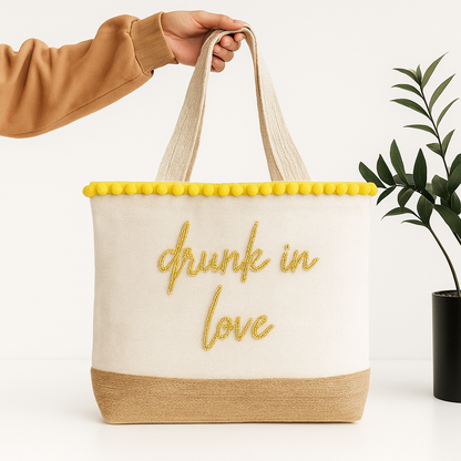 A hand holding a beige tote bag with the text "drunk in love" embroidered on it, against a white background.