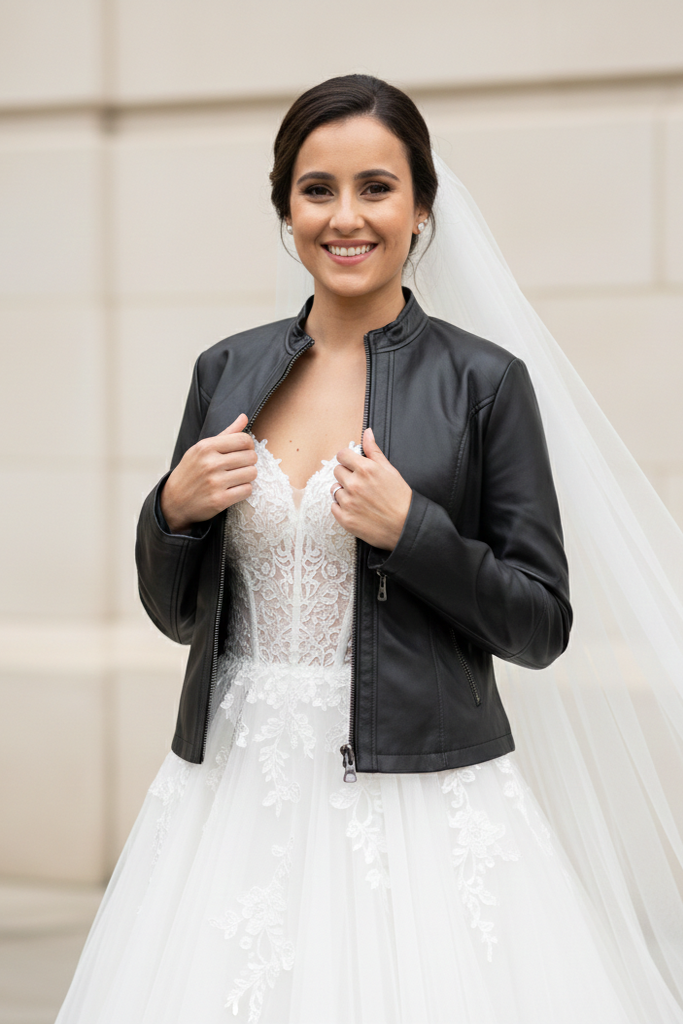 A woman in a white wedding dress with a black leather jacket and veil stands in front of a beige wall.