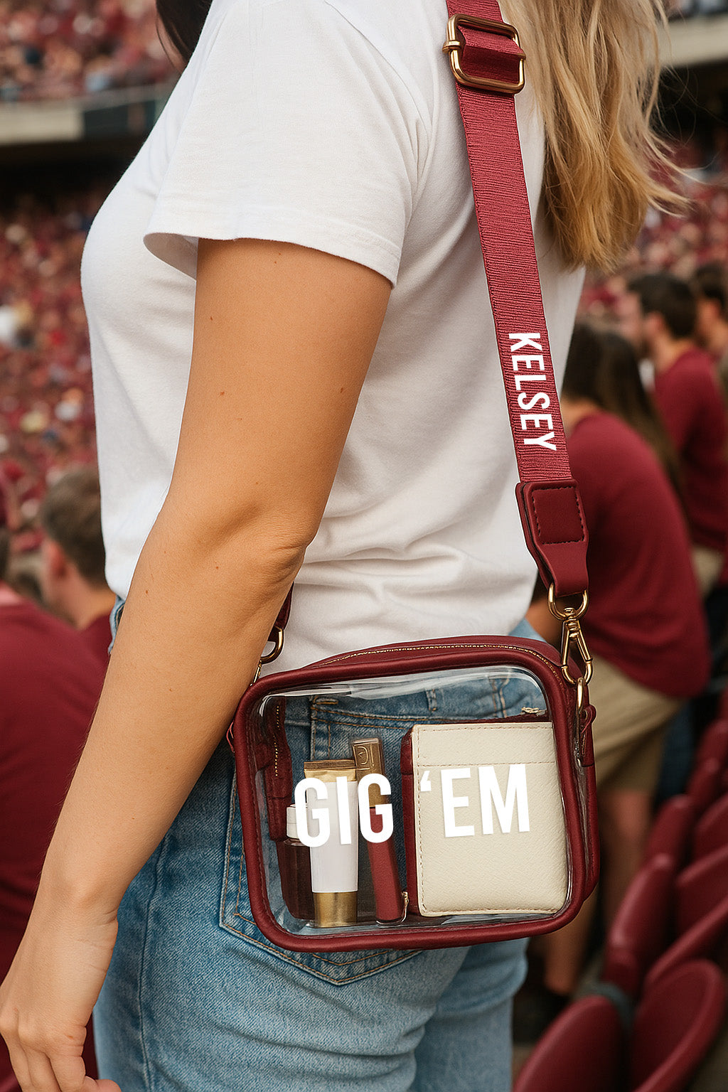 A person wearing a white t-shirt and blue jeans is holding a red purse with a clear plastic window, which has the text "GIG 'EM" printed on it.