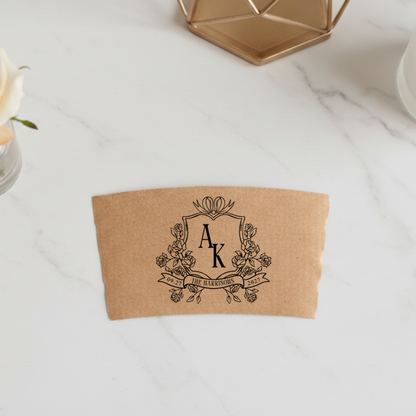 A brown paper coffee cup sleeve with a monogrammed logo printed on it, placed on a white marble surface.