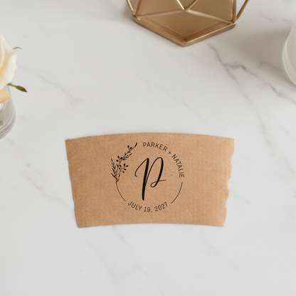 A brown paper coffee cup sleeve with a monogrammed design and text, placed on a white marble surface.