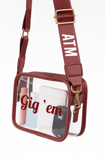 A maroon crossbody bag with a clear front pocket, displaying various items such as a wallet, keys, and a phone.