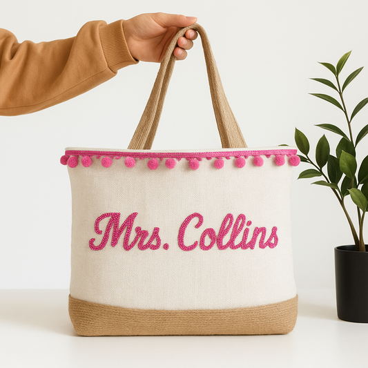 A hand holding a beige tote bag with pink pom poms and the name "Mrs. Collins" embroidered on it.