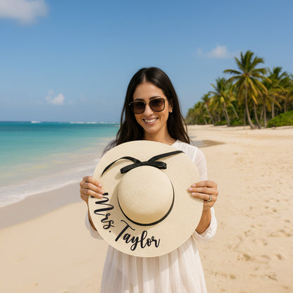 A woman wearing sunglasses and a white dress holds a large white hat with the name "Mrs. Taylor" written on it, standing on a beach with palm trees in the background.