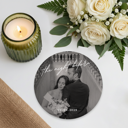 a round coaster with a black and white photograph of a couple embracing, surrounded by a bouquet of white roses and greenery.