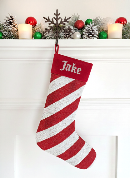 A red and white striped Christmas stocking with the name "Jake" written on it, hanging on a mantle decorated with Christmas ornaments and lights.