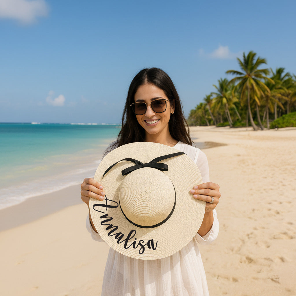 A woman wearing a white dress and sunglasses holds up a large white hat with the name "Annalisa" written on it, standing on a beach with palm trees in the background.