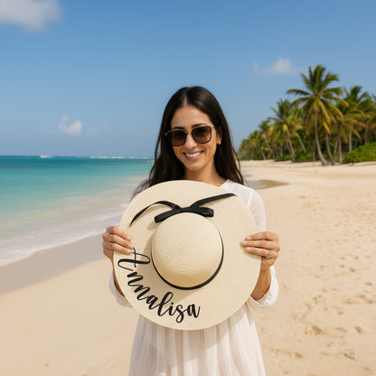 A woman wearing a white dress and sunglasses holds up a large white hat with the name "Annalisa" written on it, standing on a beach with palm trees in the background.