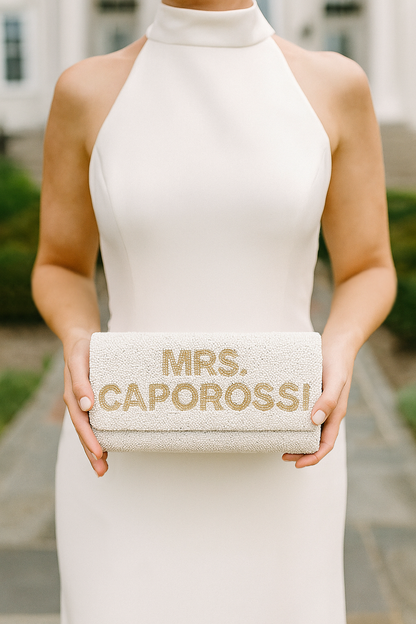 A person wearing a white dress is holding a beaded clutch with the text "MRS. CAPOROSSO" printed on it.