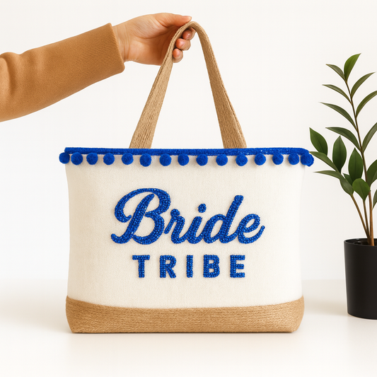 A hand holding a white tote bag with blue text and decorative pom-poms on the edges, placed next to a potted plant.