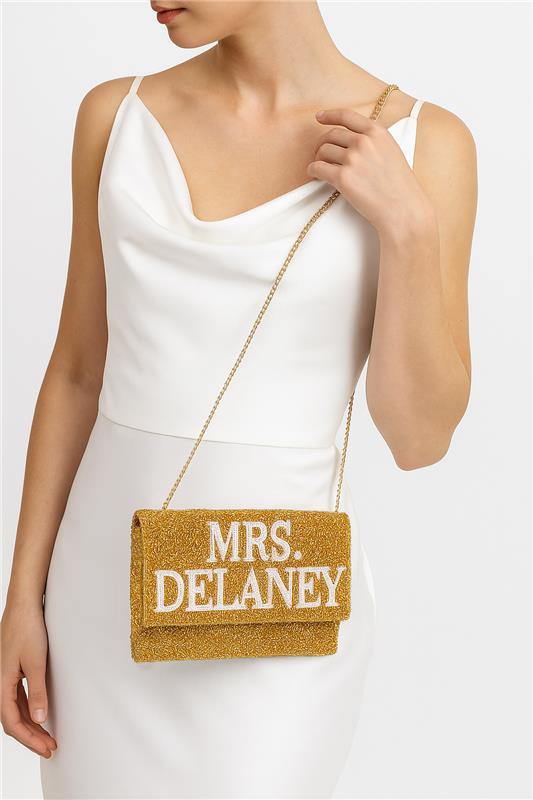 A woman wearing a white dress is holding a gold clutch purse with the text "Mrs. Delaney" printed on it.