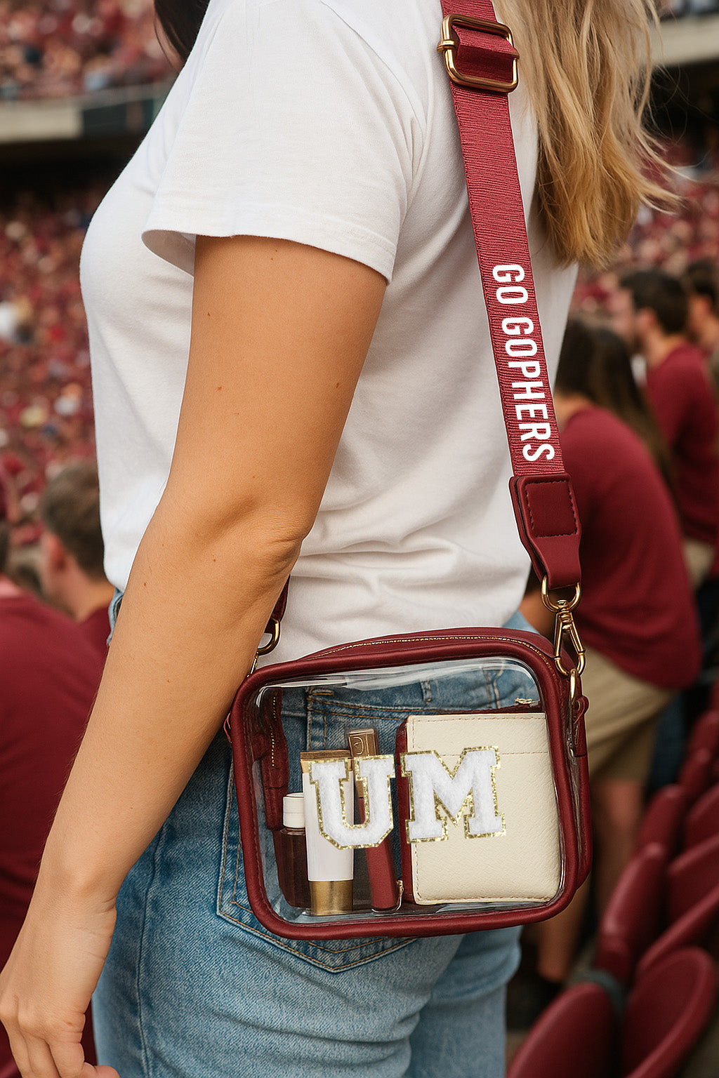 A person wearing a white t-shirt and blue jeans is holding a maroon purse with a clear plastic window, displaying the letters "UM" on it.