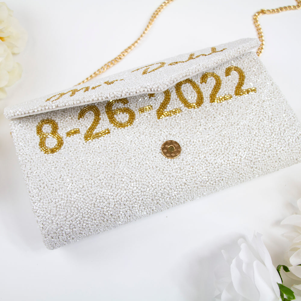 A personalized envelope bridal clutch with the option for a custom last name, meticulously beaded for an elegant touch. Measuring 10" W x 6" H with a gusset, this handbag is ideal for wedding day essentials. It can come with either a gold or silver chain, and features an interior custom date. This unique, handmade bridal clutch showcases the individuality and variations inherent in artisan crafts