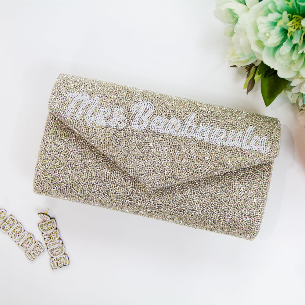 A personalized envelope bridal clutch with the option for a custom last name, meticulously beaded for an elegant touch. Measuring 10" W x 6" H with a gusset, this handbag is ideal for wedding day essentials. It can come with either a gold or silver chain, and features an interior custom date. This unique, handmade bridal clutch showcases the individuality and variations inherent in artisan crafts