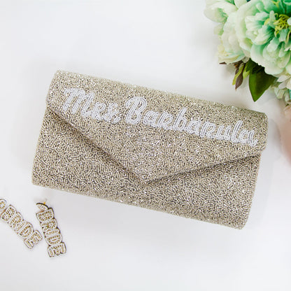 A personalized envelope bridal clutch with the option for a custom last name, meticulously beaded for an elegant touch. Measuring 10" W x 6" H with a gusset, this handbag is ideal for wedding day essentials. It can come with either a gold or silver chain, and features an interior custom date. This unique, handmade bridal clutch showcases the individuality and variations inherent in artisan crafts