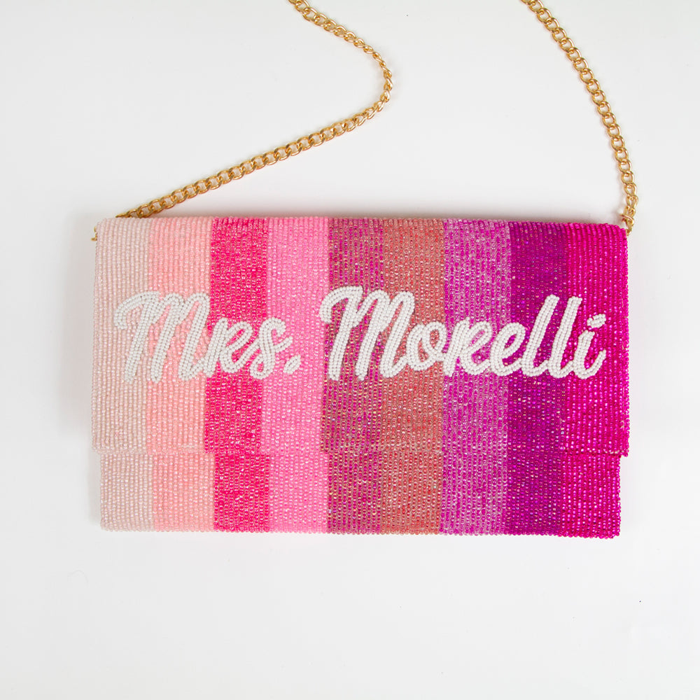 Exquisite Personalized Pink Ombre Clutch Bag, masterfully handcrafted for wedding occasions. Measuring 9.5 inches in length and 6 inches in breadth, this clutch features a gradient of pink hues, accented with white text beading. Complete with a gold or silver chain and plush velvet interior, it can be personalized with a date inside. A perfect blend of elegance and functionality, it's a distinctive accessory that adds a touch of sophistication to bridal attire.