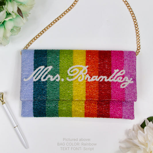 Elegant Custom Rainbow Clutch Purse, ideal for wedding occasions. Handcrafted with precision, this 9.5 inches wide by 5 inches tall clutch showcases a vibrant rainbow design complemented by white text beading. It boasts a soft velvet interior and can be adorned with a gold or silver chain. The option for a personalized date on the inside adds a unique touch, making it both a stylish and sentimental accessory for brides or as a heartfelt gift