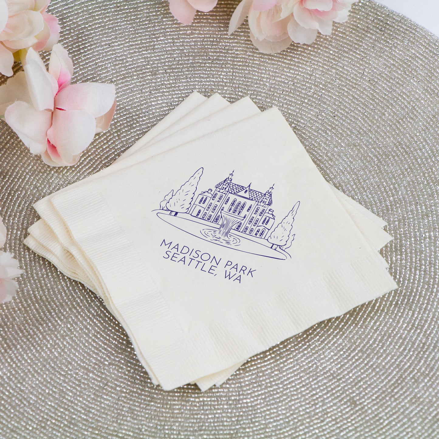 Stack of white napkins with a blue logo and text on a textured surface with pink flowers.