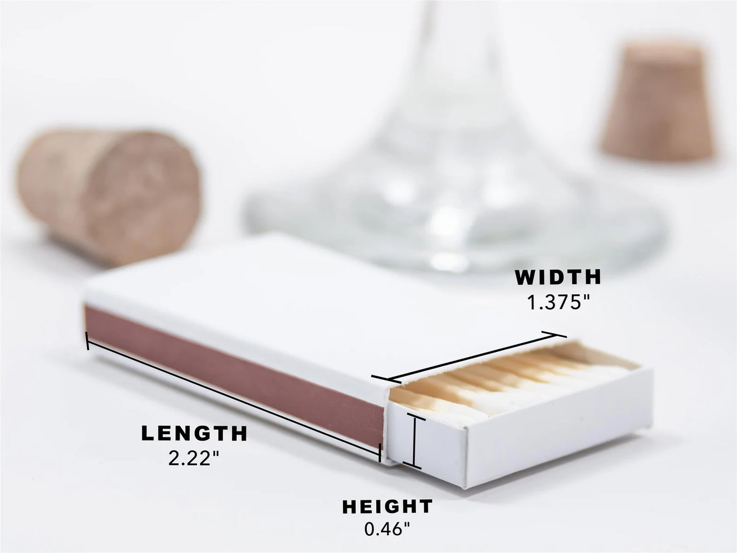a matchbox with a cork stopper and a wine glass in the background. The matchbox is described as having a length of 2.22 inches, a height of 0.46 inches, and a width of 1.375 inches.