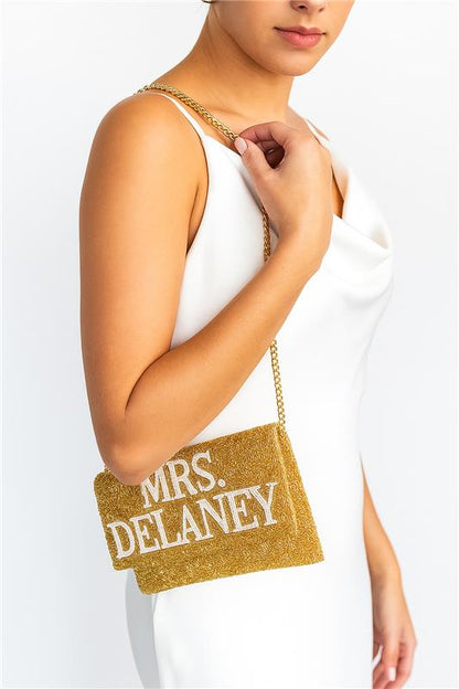 A woman wearing a white dress is holding a gold clutch purse with the text "Mrs. Delaney" printed on it.