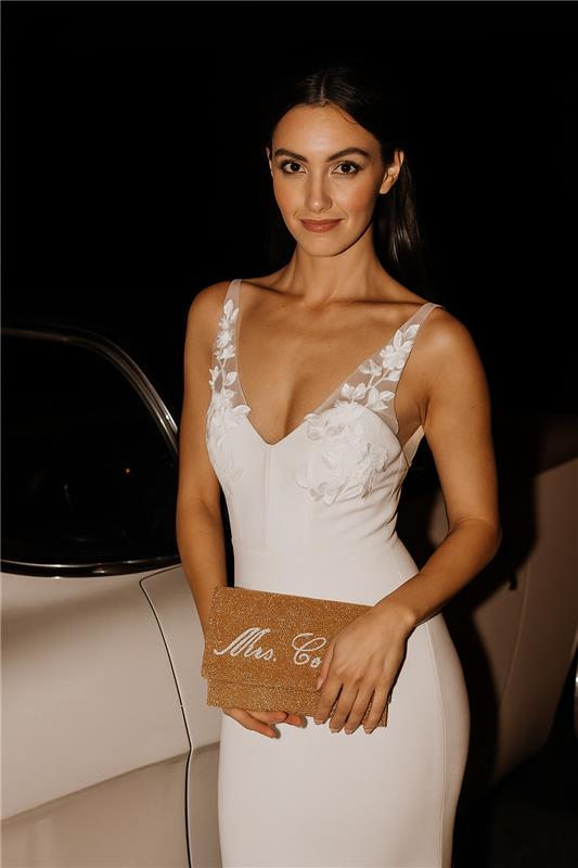 A woman in a white dress stands next to a white car, holding a gold clutch purse with the text "Mrs. Co." printed on it.