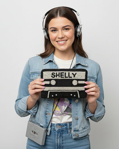 A young woman wearing headphones and a denim jacket is holding a black and white cassette tape bag with the word "SHELLY" printed on it.