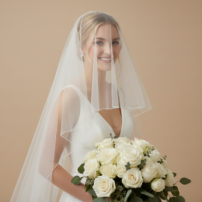A smiling bride in a white wedding dress holding a bouquet of white roses stands against a beige background.