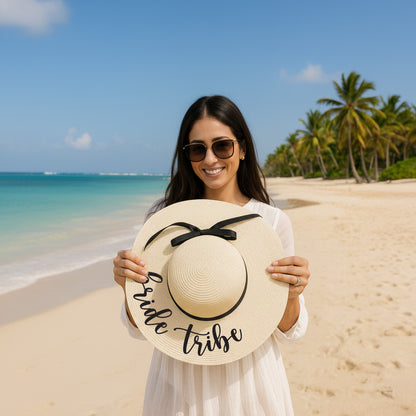 A woman in a white dress holds a large white hat with the text "Bride Tribes" on it, standing on a beach with palm trees and a clear blue sky in the background.