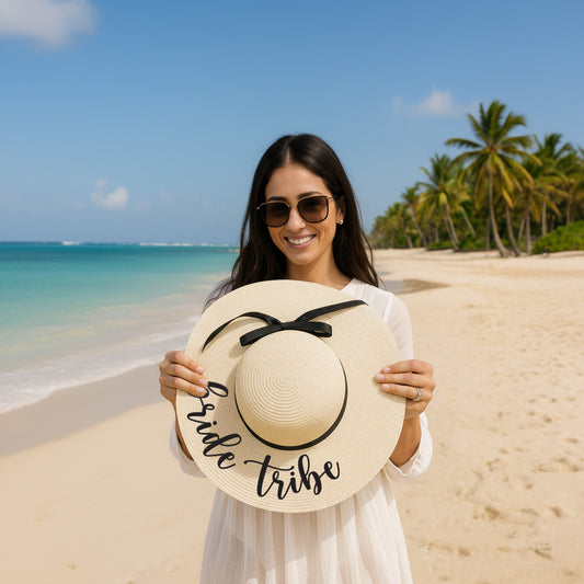 A woman in a white dress holds a large white hat with the text "Bride Tribes" on it, standing on a beach with palm trees and a clear blue sky in the background.
