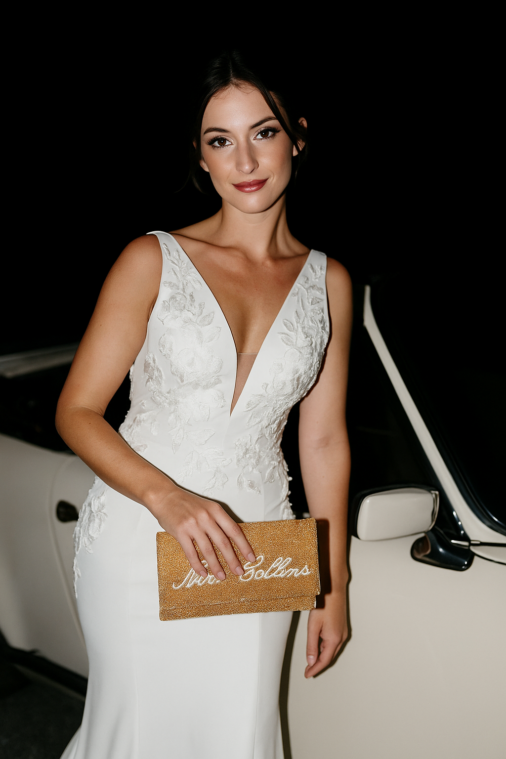 A woman in a white wedding dress stands next to a white car, holding a gold clutch purse with the text "Sophie & Gollins" on it.