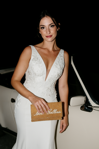 A woman in a white wedding dress stands next to a white car, holding a gold clutch purse with the text "Sophie & Gollins" on it.