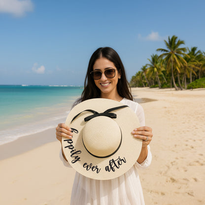 A woman in a white dress stands on a beach holding a large white hat with the text "happily ever after" written on it.
