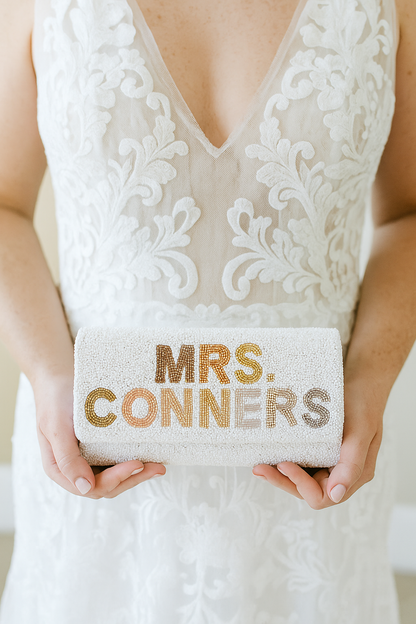 A person is holding a small white clutch purse with the text "MRS. CONNERS" printed on it.