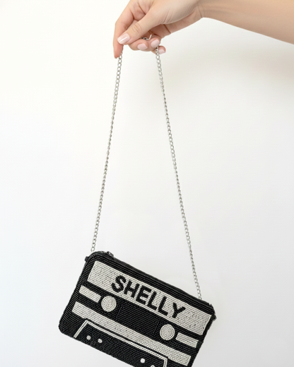 A hand holding a black and white handbag with the word "SHELLY" printed on it.