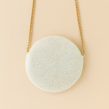 A white, round, handbag with a gold chain strap is shown against a light peach background.