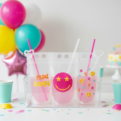 A table with three clear plastic cups filled with pink liquid, each with a straw and a smiley face design. The cups are surrounded by colorful confetti on the table.