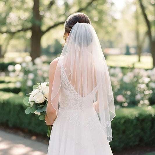 A bride in a white wedding dress holding a bouquet of white flowers stands in a garden, with a lush green hedge and trees in the background.