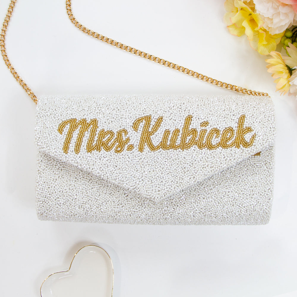 A personalized envelope bridal clutch with the option for a custom last name, meticulously beaded for an elegant touch. Measuring 10" W x 6" H with a gusset, this handbag is ideal for wedding day essentials. It can come with either a gold or silver chain, and features an interior custom date. This unique, handmade bridal clutch showcases the individuality and variations inherent in artisan crafts
