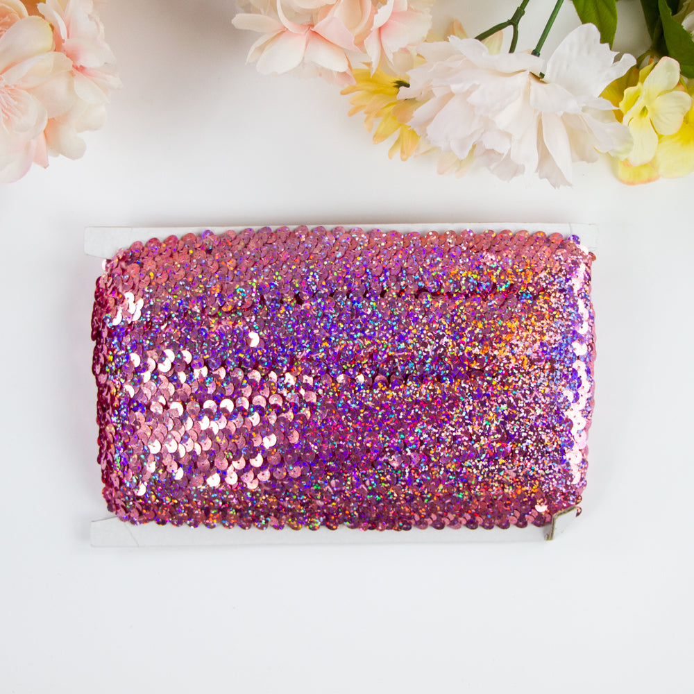 Holographic light pink stretch sequin trim roll, 1 inch wide and 11 yards in length. Ideal for embellishing t-shirts, dance wear, and home decor with its vibrant pink sparkly sequins. Easy to sew onto various fabrics for a dazzling finish