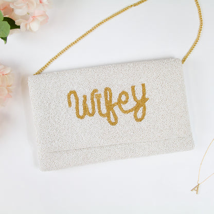 Elegant Wifey Bride Clutch Purse adorned with shimmering gold text, handcrafted with an intricate beaded design. Measuring 10.75 x 6.75 x 1 in and made of durable canvas material, it features a magnetic snap closure for security. This chic bridal purse, perfect for holding essentials, adds a sophisticated touch to any wedding ensemble. Its uniqueness is emphasized by the handcrafted nature, ensuring that each purse is a distinctive masterpiece.