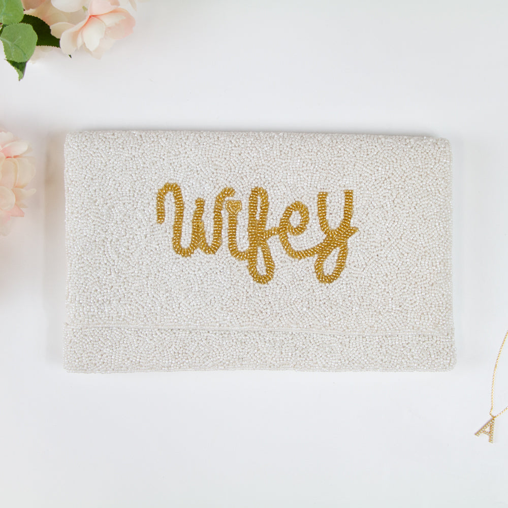 Elegant Wifey Bride Clutch Purse adorned with shimmering gold text, handcrafted with an intricate beaded design. Measuring 10.75 x 6.75 x 1 in and made of durable canvas material, it features a magnetic snap closure for security. This chic bridal purse, perfect for holding essentials, adds a sophisticated touch to any wedding ensemble. Its uniqueness is emphasized by the handcrafted nature, ensuring that each purse is a distinctive masterpiece.