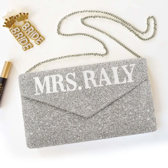 A personalized envelope bridal clutch with the option for a custom last name, meticulously beaded for an elegant touch. Measuring 10" W x 6" H with a gusset, this handbag is ideal for wedding day essentials. It can come with either a gold or silver chain, and features an interior custom date. This unique, handmade bridal clutch showcases the individuality and variations inherent in artisan crafts